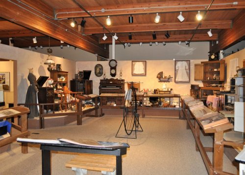 A museum interior featuring vintage furniture, clothing, and artifacts displayed in a well-lit, wooden space.