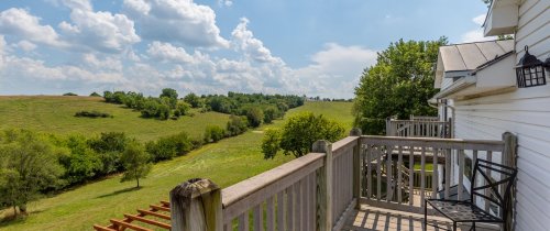 A scenic view from a balcony overlooking green fields and trees under a partly cloudy sky.