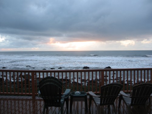 A serene ocean view at sunset, with three chairs on a wooden deck overlooking the waves and rocky shoreline.