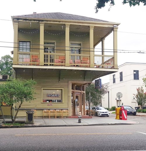 A two-story yellow building with a balcony, featuring outdoor seating and a storefront on a street corner.