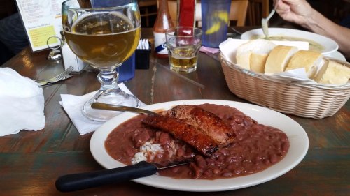 A plate of red beans and rice topped with grilled sausage, accompanied by a glass of beer and a basket of bread.