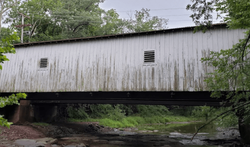 A weathered covered bridge spans a small creek, surrounded by lush greenery and trees.