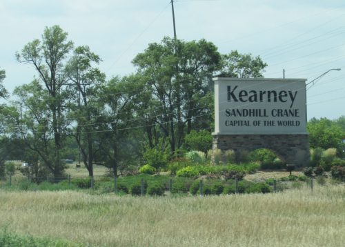 Sign for Kearney, labeled "Sandhill Crane Capital of the World," surrounded by trees and grassy fields.
