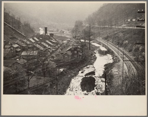 A black-and-white photo of a small town by a river, with houses, trees, and train tracks in a foggy landscape.