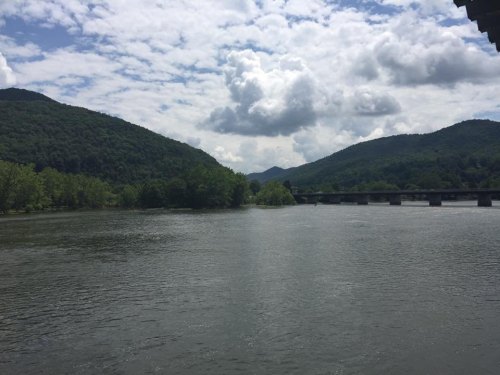 A serene river flows between lush green mountains under a partly cloudy sky, with a bridge in the distance.