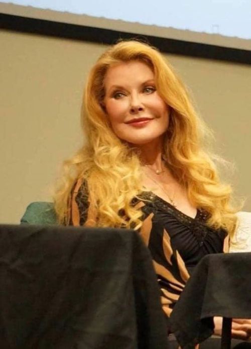 A woman with long, wavy blonde hair smiles while seated at a table, wearing a black and gold outfit.