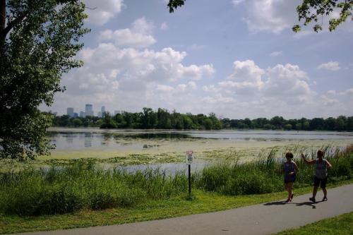 Two people jog along a path by a lake, with lush greenery and a city skyline in the background under a cloudy sky.