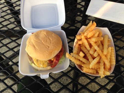 A cheeseburger with lettuce and bacon in a takeout container, alongside a serving of crinkle-cut fries on a table.