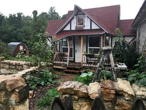 A charming cottage with a wooden porch, surrounded by lush greenery and a stone fence.