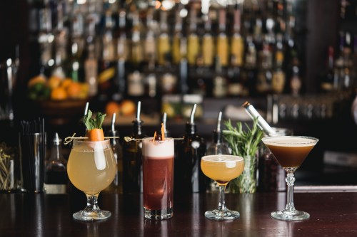 Four colorful cocktails on a bar counter, with a blurred background of bottles and fresh fruit.