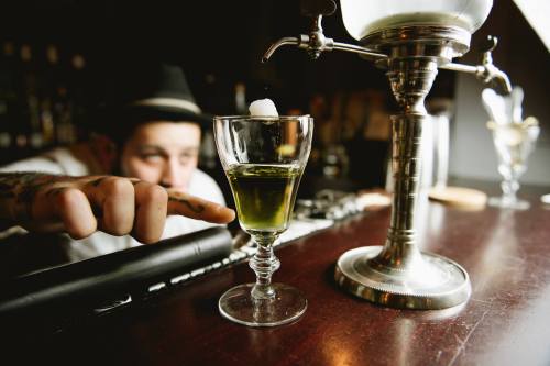 A bartender points at a green cocktail in a glass, with a sugar cube on top, set on a polished bar counter.