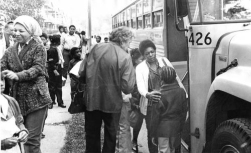 A group of people, including children and adults, gather near a school bus in a neighborhood setting.