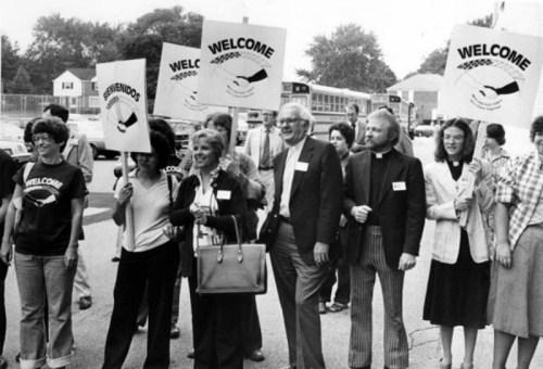 Black and white photo of a diverse group of people holding "Welcome" signs at a rally or event.
