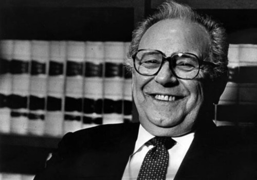 A smiling older man with glasses, wearing a suit and tie, sits in front of shelves filled with books.