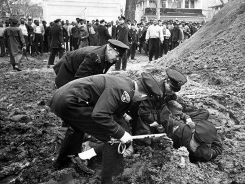 Police officers assist a person on the ground in a crowded outdoor scene, with spectators watching nearby.