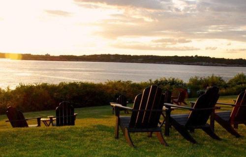 Adirondack chairs on a grassy shore overlooking a calm lake at sunset, with clouds in the sky.