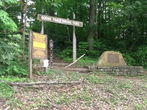 Sign for Boone Trace Hiking Trail with informational plaques and a stone monument, surrounded by greenery.