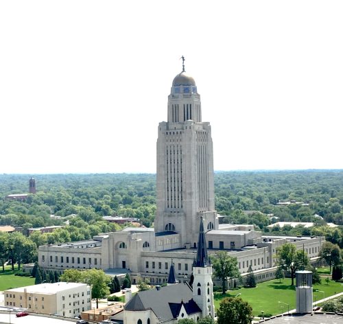A tall, art deco building with a dome, surrounded by green trees and a clear sky, likely a government structure.