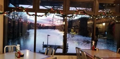 A cozy restaurant interior with festive decorations, overlooking a serene lake at sunset through large windows.