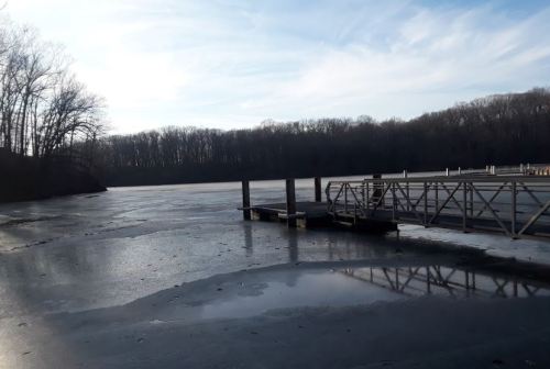 A serene winter scene of a frozen lake with a wooden dock and bare trees in the background under a clear sky.