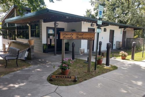 Exterior of a family restaurant with a sign, flower pots, and a welcoming path. Trees and benches are nearby.