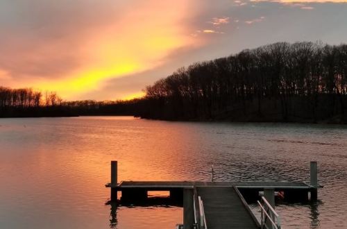 A serene sunset over a calm lake, with a wooden dock extending into the water and trees silhouetted in the background.