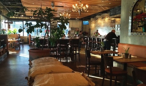 A cozy restaurant interior with wooden decor, plants, and patrons dining at tables. Sunlight streams through the windows.