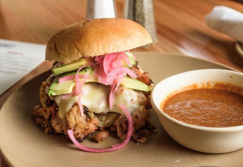 A delicious sandwich with grilled chicken, avocado, pickled onions, and melted cheese, served with a side of sauce.