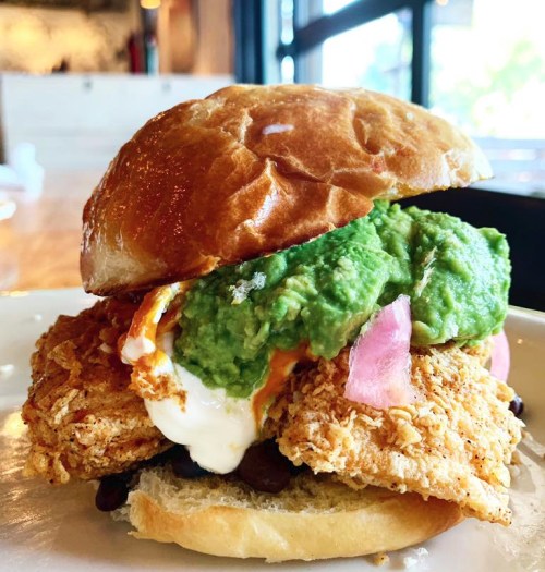 A crispy fried chicken sandwich topped with guacamole, pickled onions, and creamy sauce on a toasted bun.