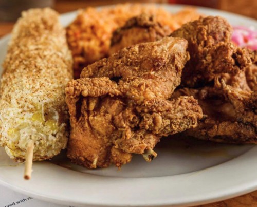 A plate featuring crispy fried chicken, a corn on the cob, and a side of rice, garnished with pink onions.