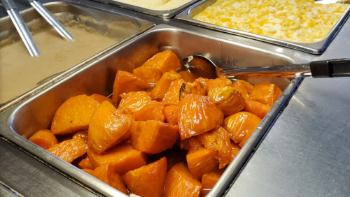 A tray of glazed orange sweet potatoes, with a serving spoon, next to trays of other dishes.