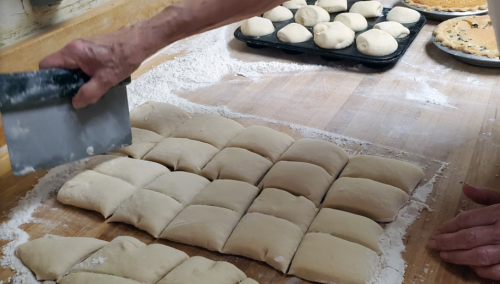 A hand using a bench scraper to cut dough into squares on a floured wooden surface, with dough balls in the background.