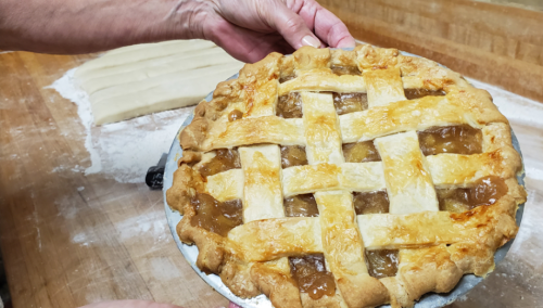 A person holds a freshly baked apple pie with a golden lattice crust, set on a wooden countertop.