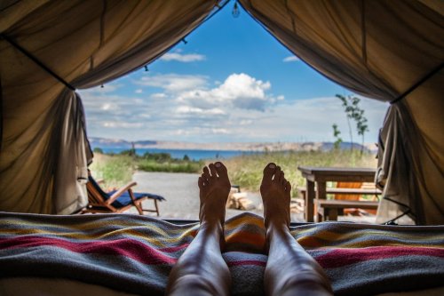 A view from inside a tent, showing bare feet on a bed with a scenic landscape and blue sky outside.