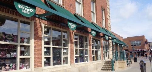 A brick building with large windows displaying antiques, featuring green awnings and a sign that reads "Antiques."
