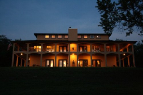 A large, two-story house illuminated at dusk, featuring multiple balconies and a grassy foreground.