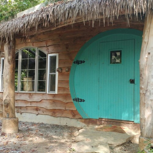 A rustic wooden cabin with a round turquoise door and large windows, surrounded by greenery.