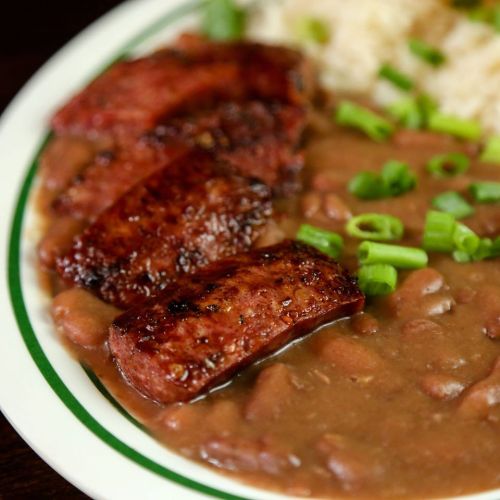 A plate of sliced, grilled sausage served with brown beans, rice, and garnished with chopped green onions.