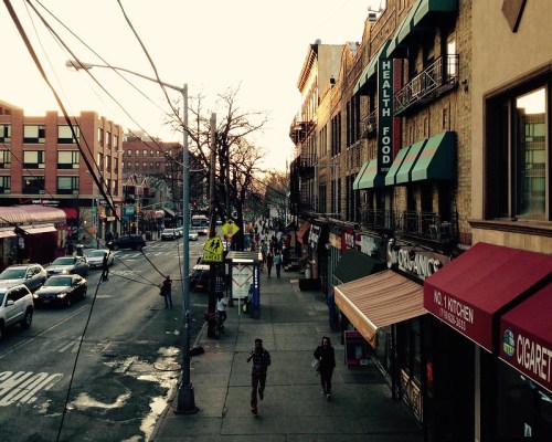 A bustling street scene at sunset, featuring shops, pedestrians, and cars along a city sidewalk.