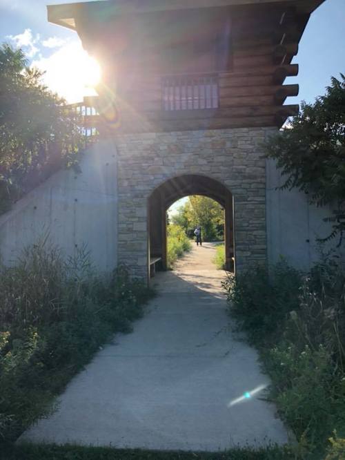 A stone archway leads to a path surrounded by greenery, with sunlight shining through a building above.