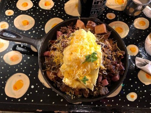 A cast iron skillet filled with a hearty breakfast of meats, cheese, and a fluffy omelet, set on a colorful table.