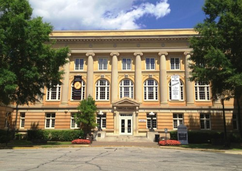 Historic building with a grand entrance, large windows, and decorative columns, surrounded by trees and flowers.