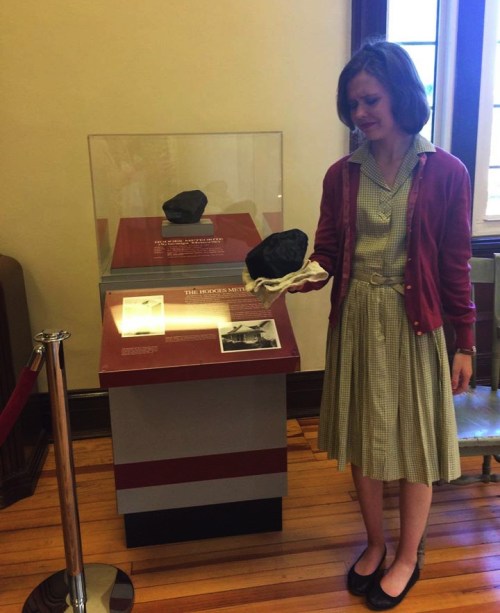 A girl in a vintage dress stands near a display case holding a black object, looking emotional in a museum setting.
