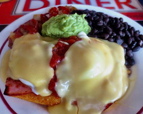 A plate of eggs Benedict topped with creamy sauce, guacamole, black beans, and salsa on a diner-style background.