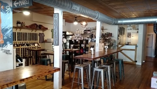 A cozy interior of a market with wooden tables, metal stools, and shelves stocked with various products.