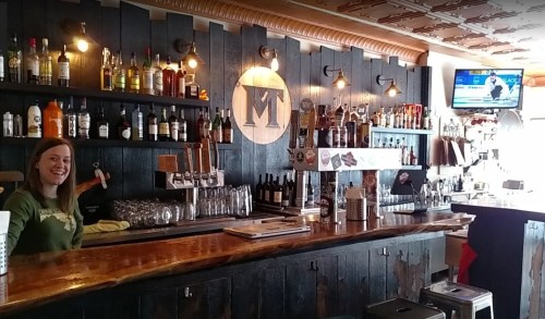 A bartender smiles behind a wooden bar stocked with various bottles and a TV showing a sports game in the background.
