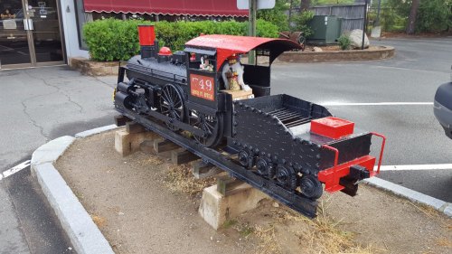 A miniature black and red steam locomotive model displayed on wooden pallets in a parking area.