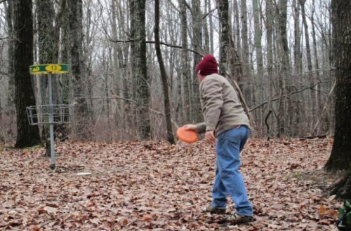 A person in a brown jacket and red beanie throws a disc towards a disc golf basket in a wooded area covered in leaves.