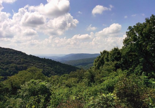A scenic view of rolling green hills under a partly cloudy sky, with distant mountains visible in the background.