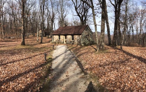 A stone cabin surrounded by bare trees and fallen leaves, with a gravel path leading to it.
